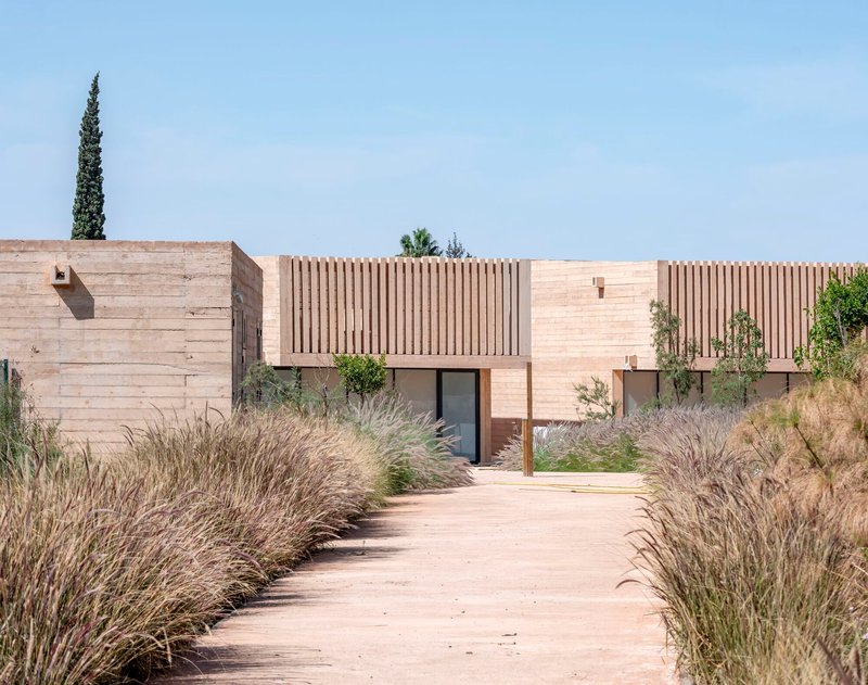 Entrance pathway bordered by wild grasses leading to rammed earth walls and vertical timber screens