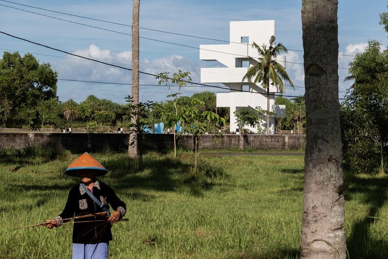 White stacked volumes rising behind grassy field with farmer in conical hat and palm trunks