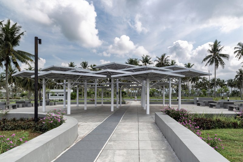 Open pavilion with folded white steel roof and circular columns framed by coconut palms