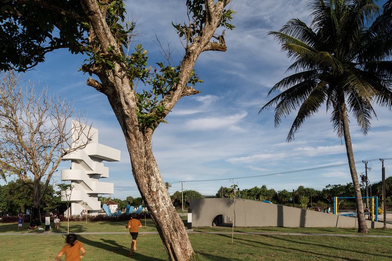 Park visitors walking past the white tower and curved concrete wall beneath palm trees