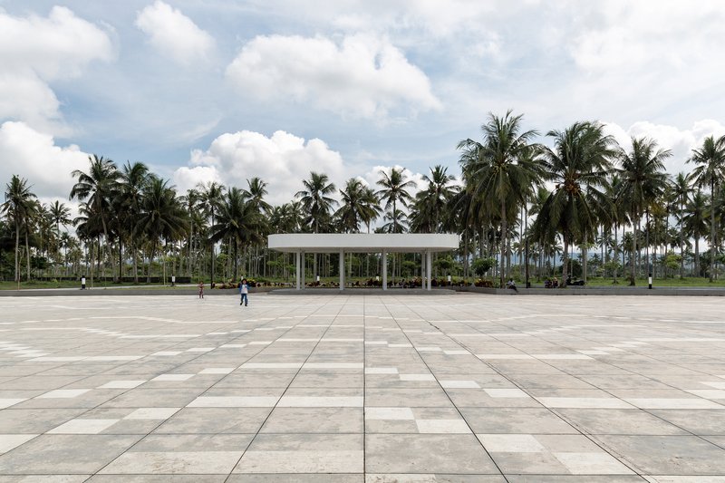 Wide paved plaza with large-format stone tiles leading to the slender-columned pavilion under a cloudy sky
