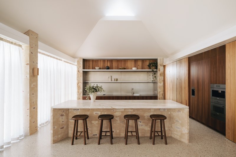 Kitchen island clad in sandstone blocks with four timber stools beneath a vaulted white ceiling