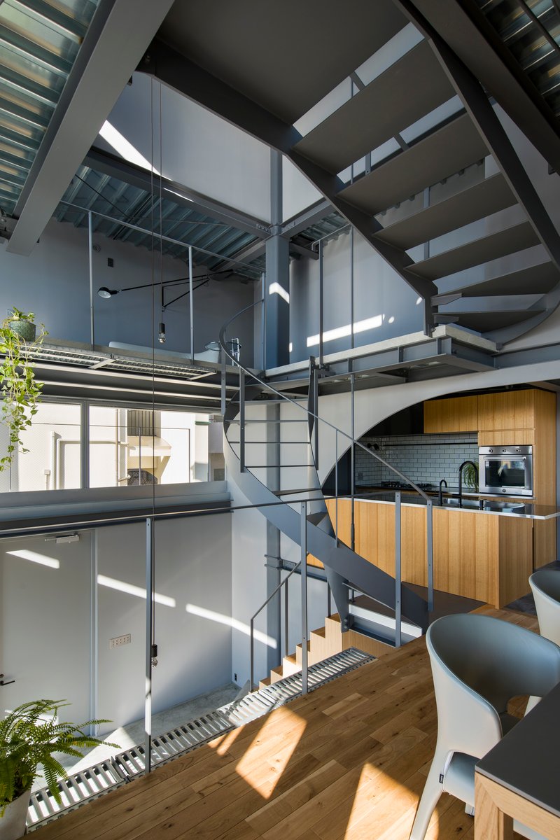 Central spiral stair winding through three floors with timber kitchen cabinetry and steel catwalks illuminated by shafts of sunlight