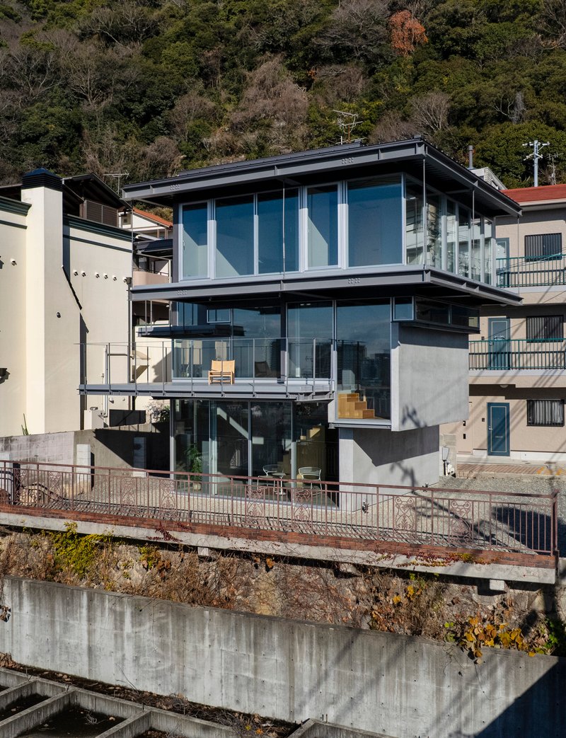 Three-story glass and concrete facade stacked on a hillside retaining wall with neighboring houses and wooded slope behind