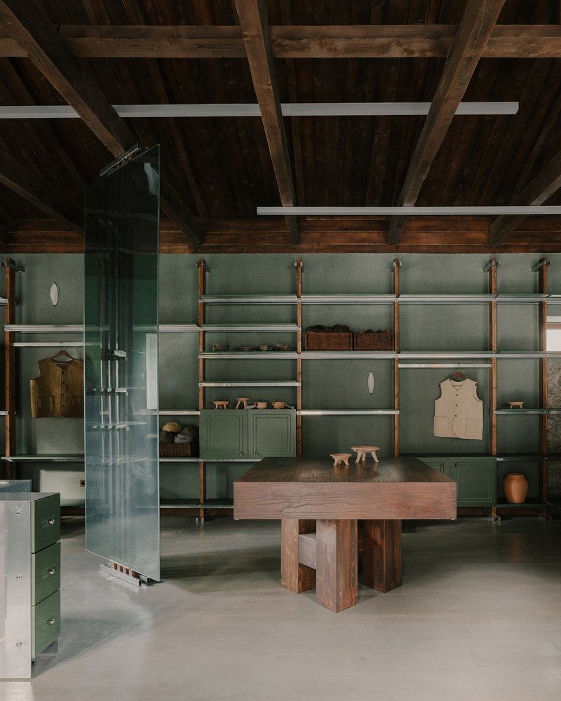 Sage green wall-mounted shelving with timber and metal supports above a solid timber table and polished concrete floor