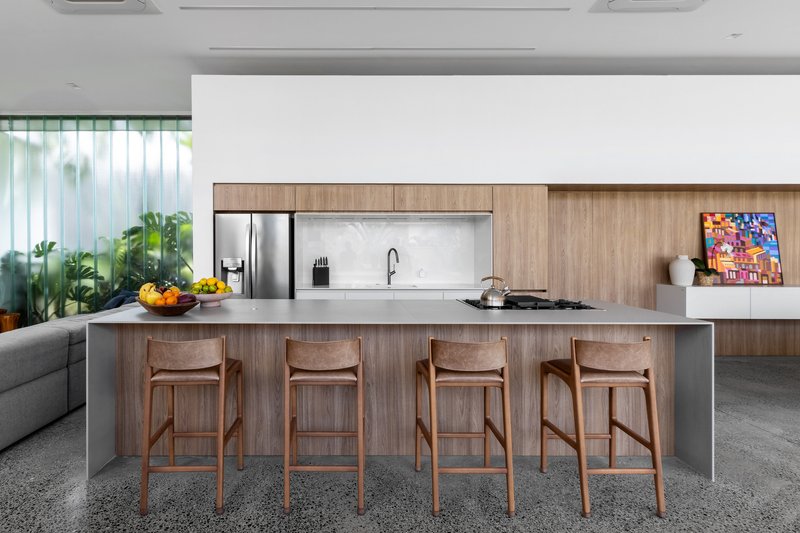 Kitchen island with timber bar stools and walnut cabinetry beside a planted internal courtyard behind glass