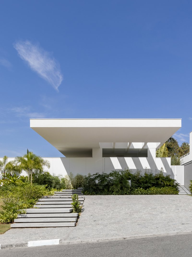 Street view of white cantilevered upper volume over stepped entrance path with palms and tropical planting