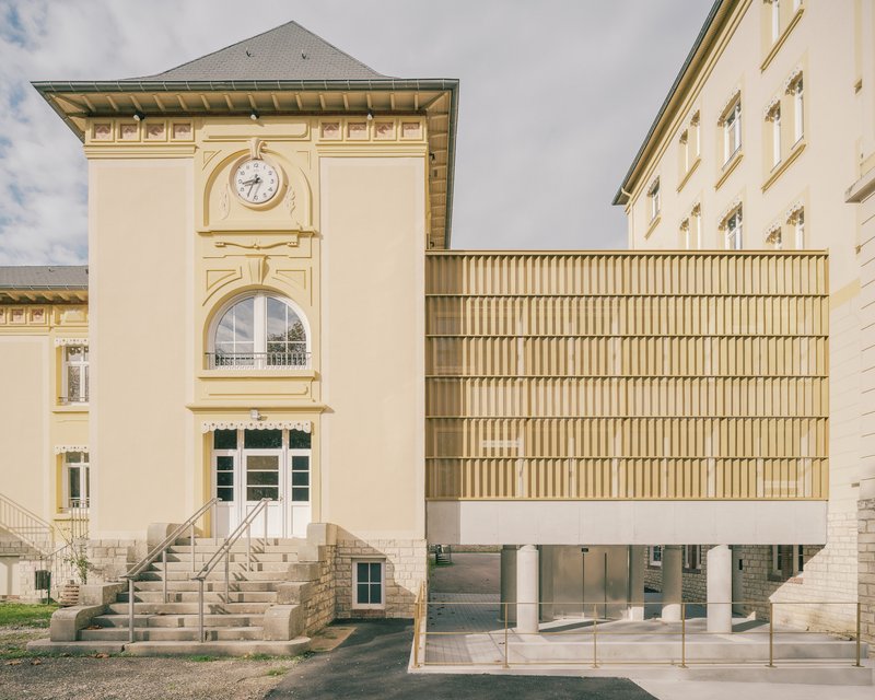 Vertical louvered timber facade on pilotis juxtaposed with a pale yellow clock tower and stone stairs