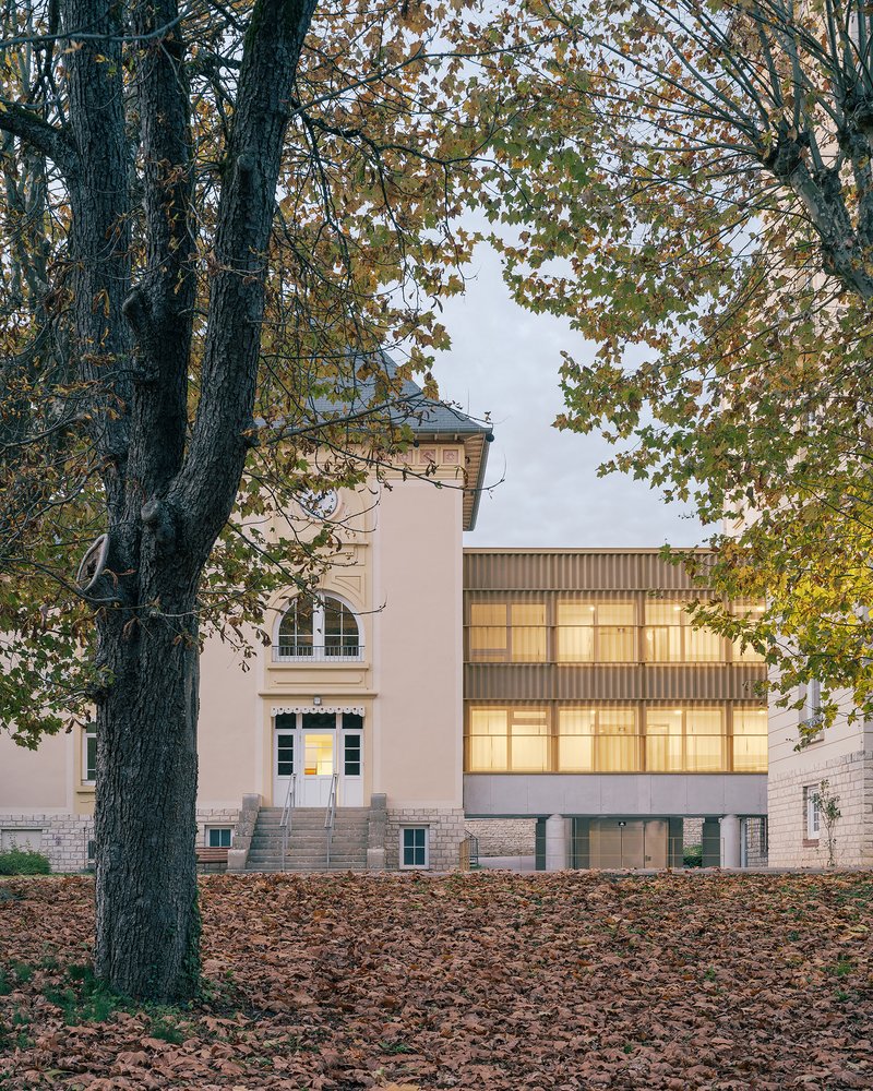 Illuminated corrugated metal facade addition between two pale plaster buildings framed by autumn foliage