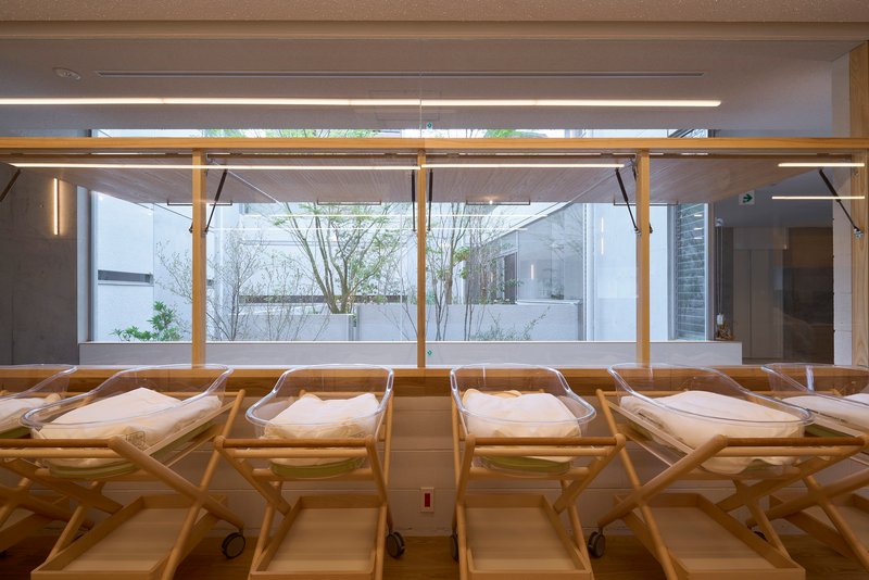Row of timber-framed infant bassinets overlooking an interior courtyard with trees through glazed wall