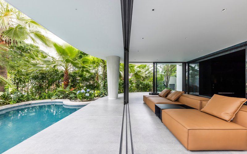 Open living pavilion with tan sectional sofa overlooking a pool and planted garden beds