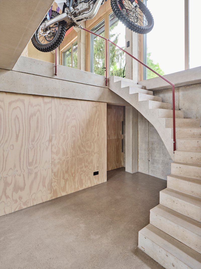 Concrete staircase with red steel handrail and plywood storage beneath displaying suspended motorcycle overhead