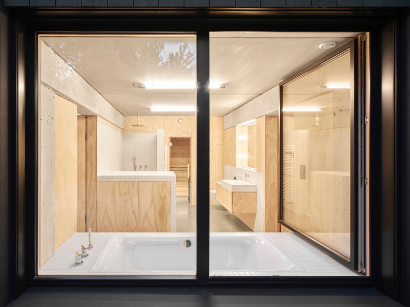 Night view through black metal window frames into bathroom with white tub and plywood cabinetry