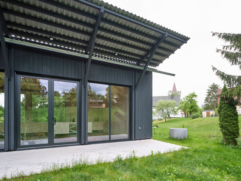 Dark timber-clad pavilion with large glass doors and corrugated metal canopy facing a grassy lawn