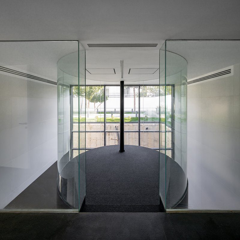 Glass-walled bridge with curved railings and central column overlooking a terraced garden courtyard
