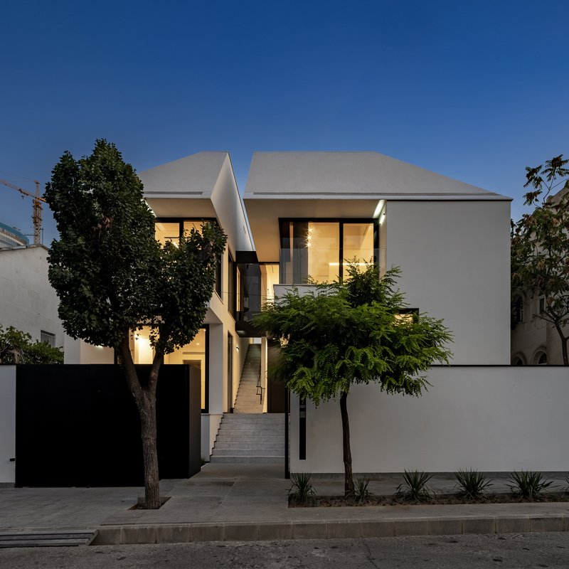 Street view of white stucco facades with illuminated windows at dusk flanked by mature trees