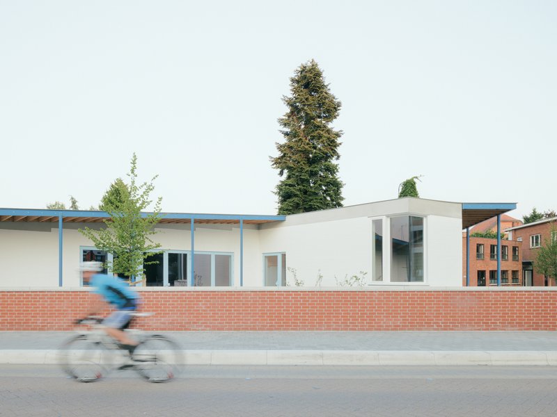 Street facade with brick base wall and white volumes under a curving roof as a cyclist passes