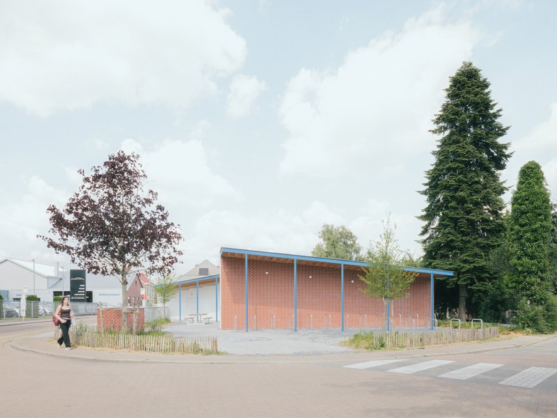 Street view of the brick-clad volume with blue steel canopy and planted low fence under a cloudy sky