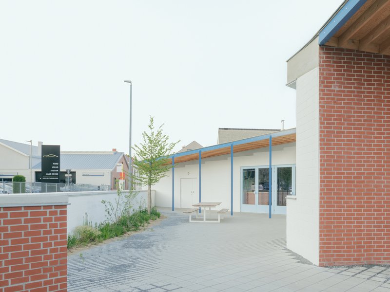 Paved courtyard entry with timber-and-steel canopy framed by two brick volumes and young trees