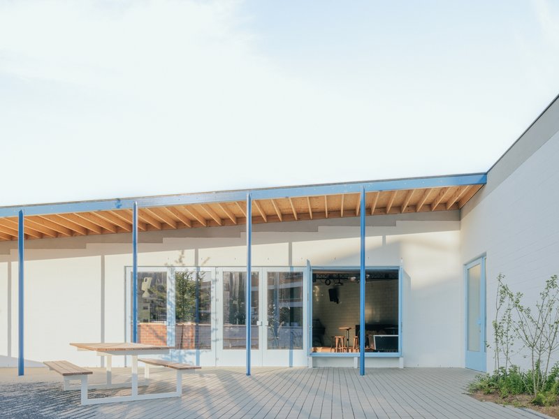 Covered terrace with exposed timber rafters and blue steel columns facing a paved courtyard and planted beds