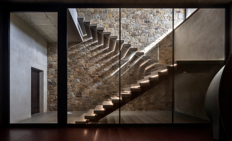 Floating steel staircase against an exposed stone wall illuminated by natural light from above