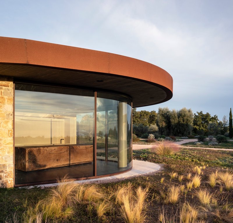 Curved glass facade with corten steel canopy and stone wall flanked by native grasses at dusk