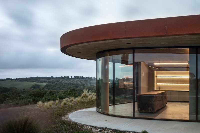 Curved glass walls beneath a corten steel overhang opening to a terrace with views of distant orchards
