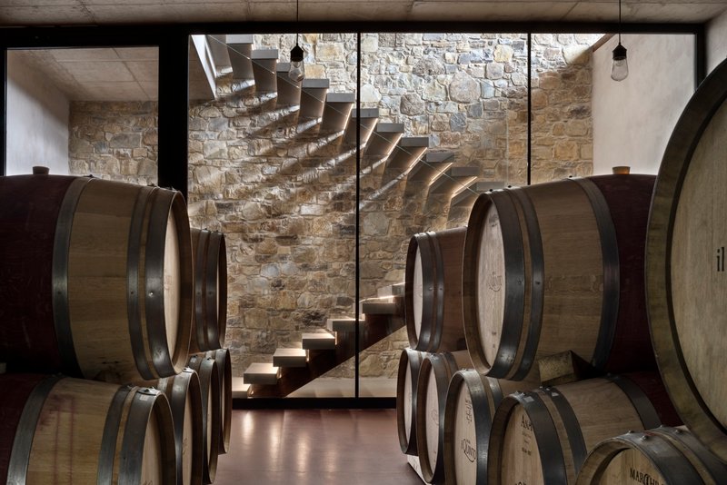 Cantilevered concrete staircase descending past stone walls and oak wine barrels