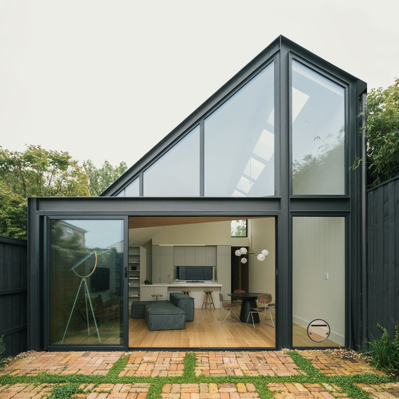 Rear garden elevation with angled glazing and black steel frames opening onto a brick and grass courtyard