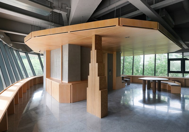 Interior with light wood pavilion structure and curved timber columns beneath exposed concrete beams and slanted glazing