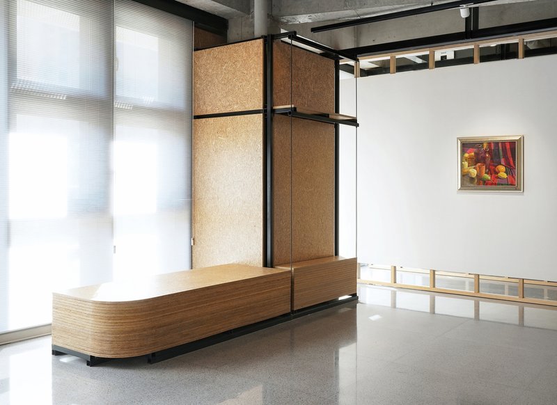 Cork-clad partition wall with black steel framing beside a curved timber desk and translucent windows