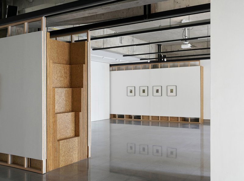 Gallery space with movable white partition panels and cork-lined recesses beneath an exposed concrete ceiling