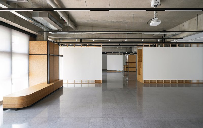 Open exhibition space with timber bench seating and white movable display walls under steel structural beams