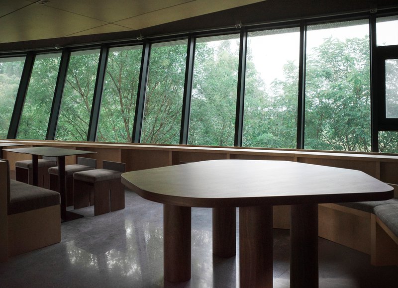 Interior dining space with curved timber table and angled window wall overlooking dense green foliage