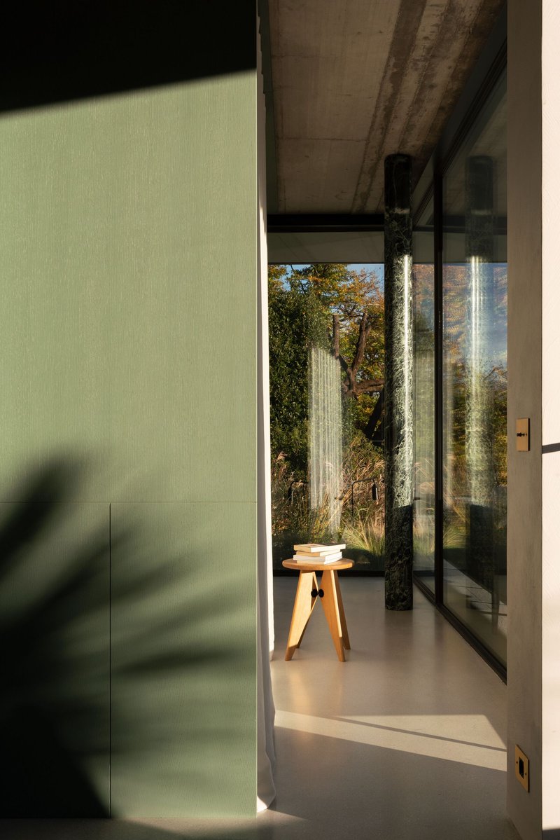 Threshold view from sage green wall to glazed corridor with timber stool and dappled sunlight