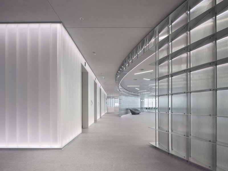 Curved corridor with translucent channel glass wall and backlit vertical panels along gray carpet floor