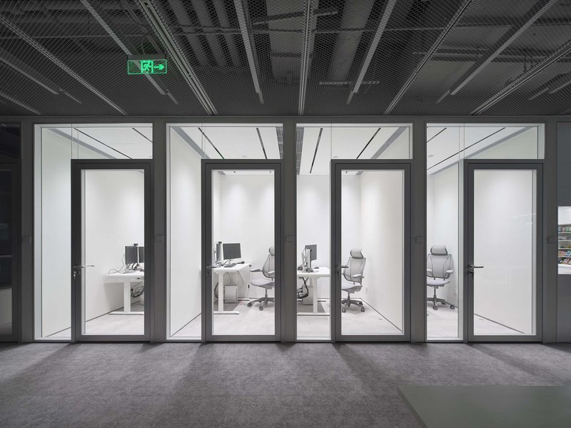 Four glass-walled office pods viewed from a corridor with exposed black ceiling and emergency exit sign