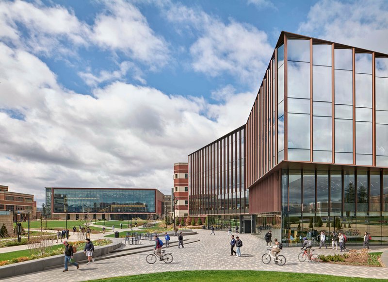 Campus plaza with paved walkways and planted beds linking vertically striped facade volumes under daytime clouds