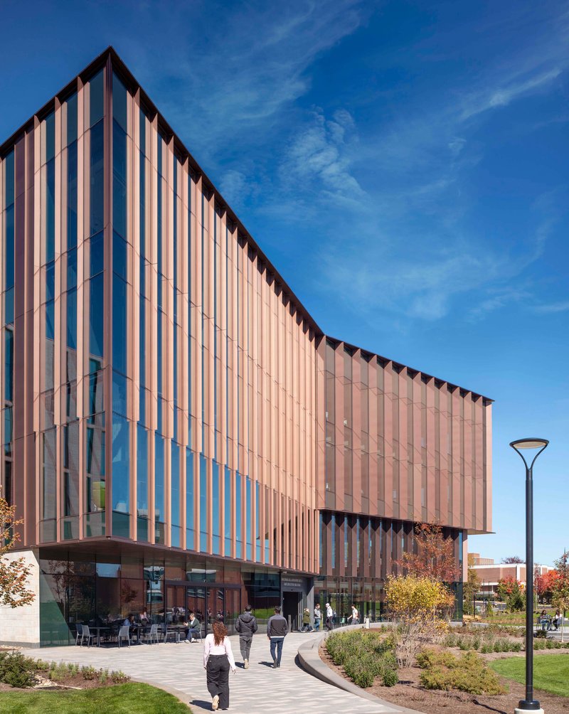 Curved facade with vertical metal fins rising above landscaped plaza in afternoon light