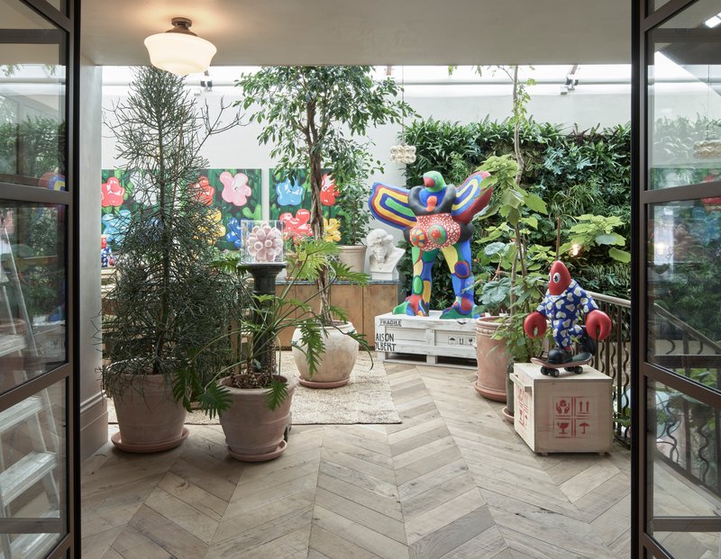 Interior courtyard with herringbone wood floor, potted plants, and colorful sculptures among steel-framed glazing