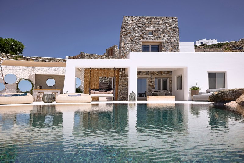 Pool deck with circular porthole screens in white pergola facing stone tower volume under clear sky
