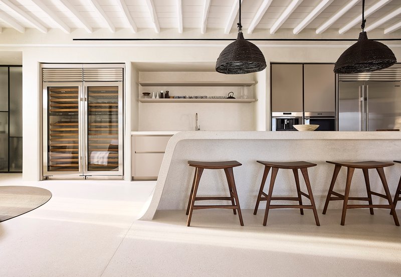 Kitchen island with curved stone edge, wooden stools, and black woven pendant lights beneath white ceiling beams