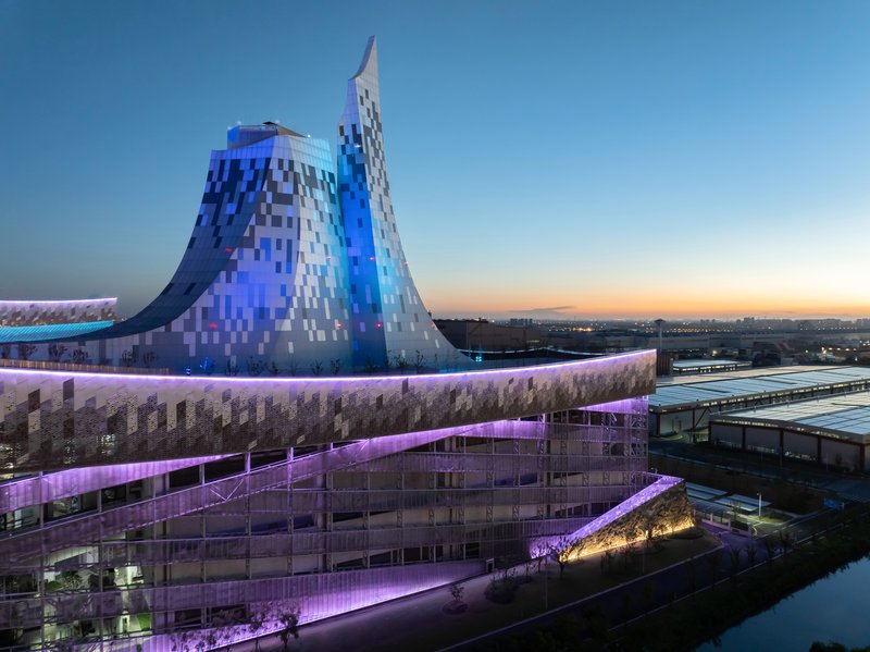 The pixelated tower with blue lighting rising above the terraced volumes at twilight