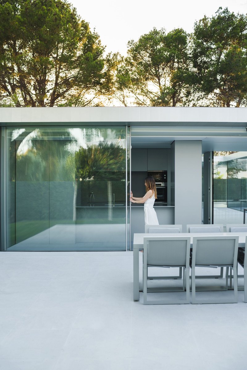 Outdoor dining terrace with glass sliding walls opening to kitchen and surrounding trees at dusk