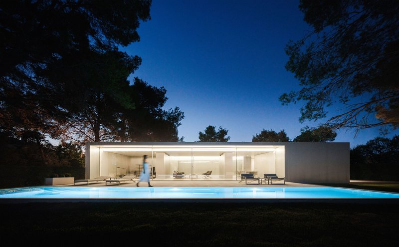 Illuminated glass facade and glowing pool terrace with a figure walking at twilight