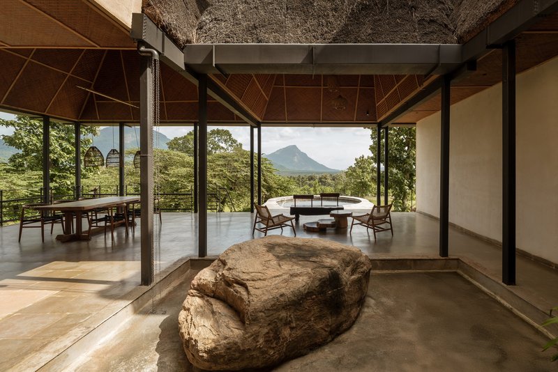 Open interior with polished concrete floor, exposed steel columns, thatch overhead, and a large weathered boulder at center