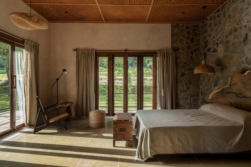 Bedroom with woven bamboo ceiling, stone accent wall, and morning sunlight streaming through timber French doors