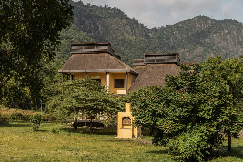 Two-story pavilion with thatched roof and ventilation cupolas set in a lawn among trees with mountains beyond