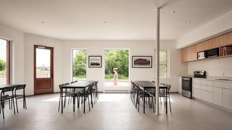 Communal dining room with large picture windows framing green foliage and a figure passing outside