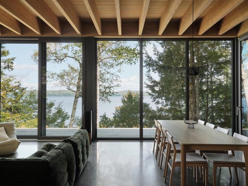 Dining and living area beneath exposed timber rafters overlooking a lake through floor-to-ceiling glazing